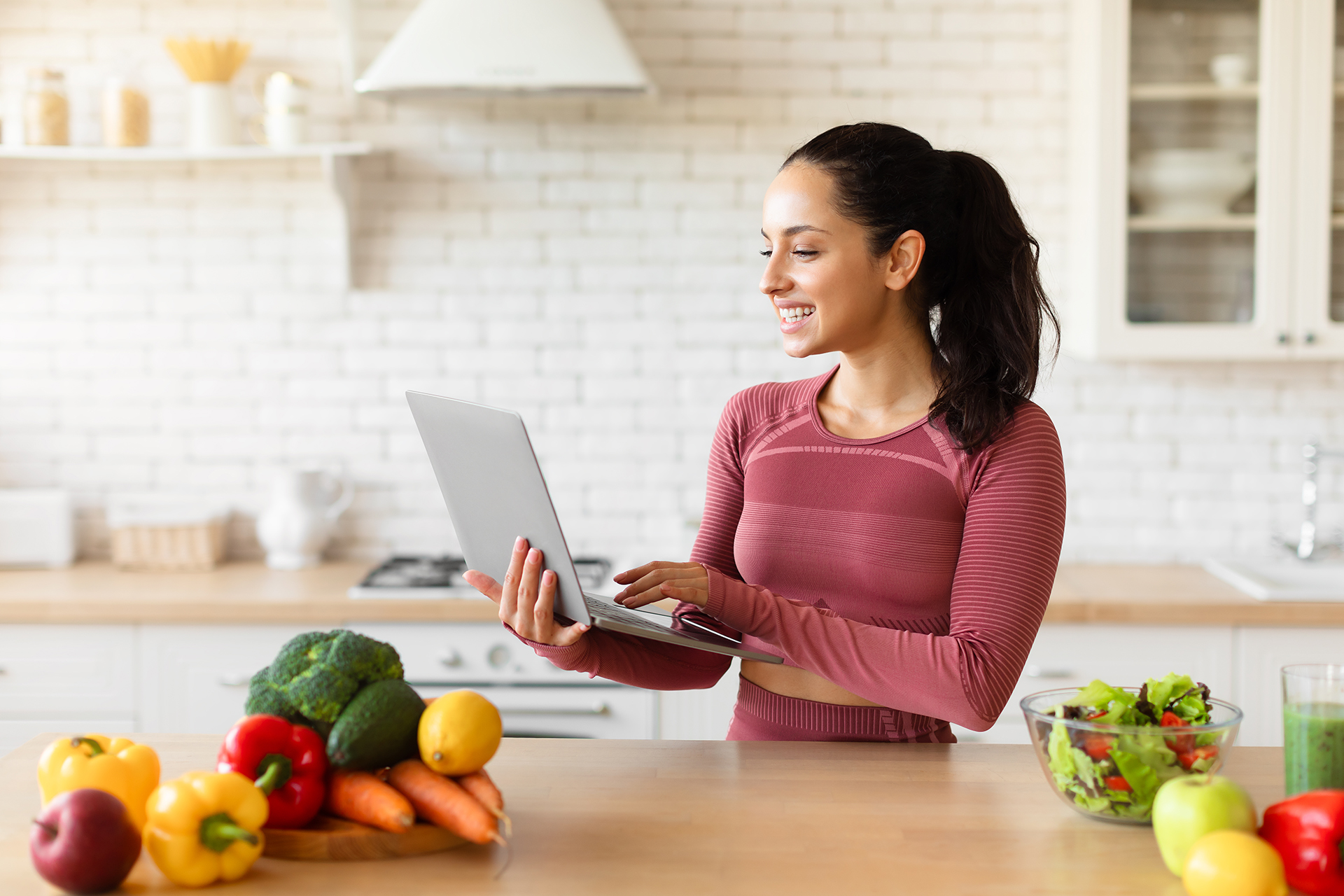 Fit lady using laptop researching healthy nutrition advice and browsing nutritious recipes for slimming, planning menu online standing in modern kitchen, wearing activewear. Weight loss concept